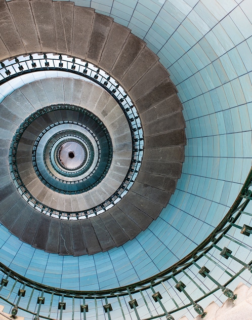 Vue plongeante d'un escalier en colimaçon en pierre grise avec rampe noire et murs bleus. Forme spirale hypnotique.