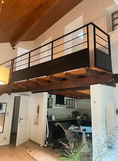 Mezzanine moderne avec balustrade noire, poutres en bois apparentes, et espace de vie ouvert sur une cuisine en contrebas.