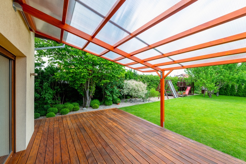 Terrasse bois moderne couverte d'un toit transparent adossée à une maison, avec jardin verdoyant et aire de jeux enfants.