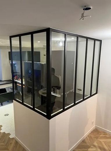 Verrière d'intérieur noire sur muret blanc, séparant une cuisine moderne avec frigo. Sol parquet et carrelage hexagonal.